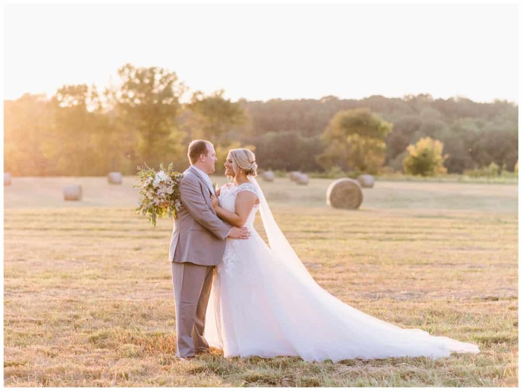 Full body photo of the bride and groom during golden hour in a hay field bails of hay in background