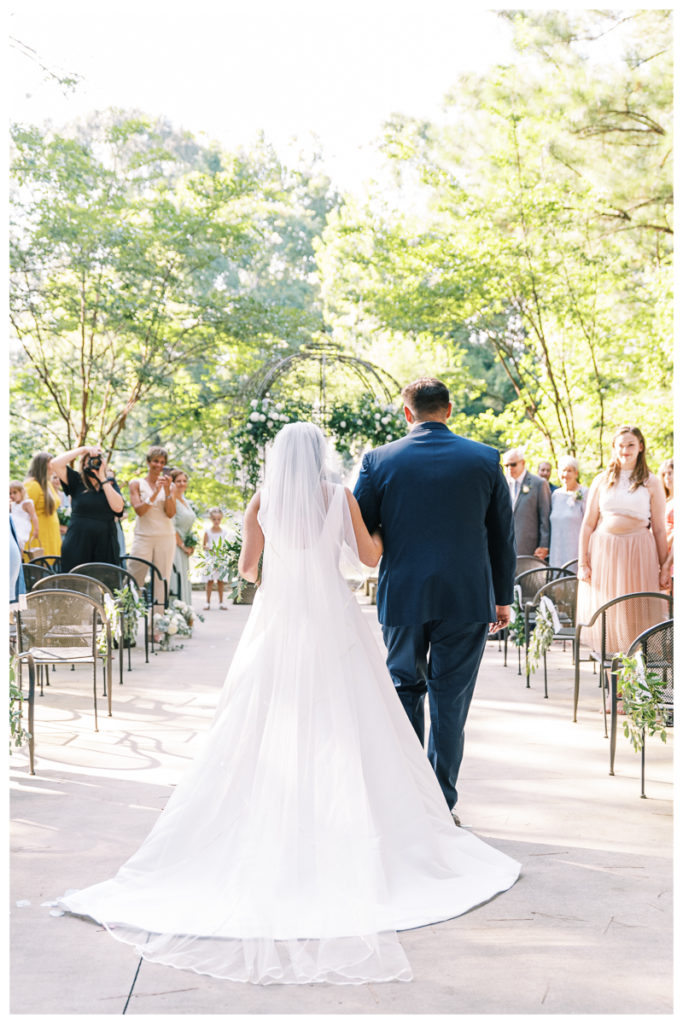 A bride walks down the aisle in an outdoor wedding ceremony with bright greenery and a flower arch